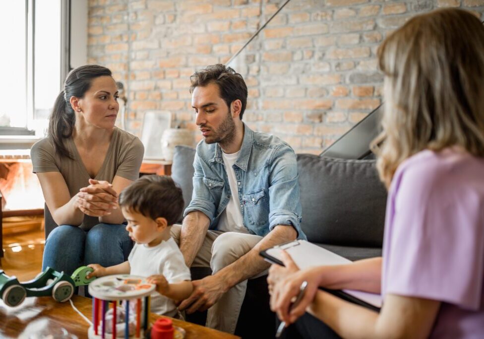 Family discussing with a counselor, toddler playing.